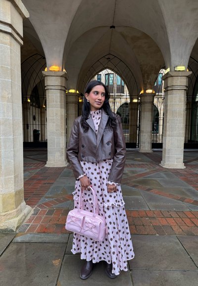 Mujer con chaqueta de cuero marrón y vestido de lunares sosteniendo un bolso rosa, de pie bajo columnas de piedra arqueadas con suelo de baldosas.