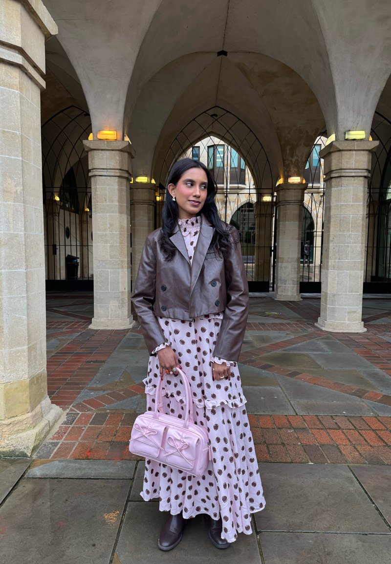 Γυναίκα με καφέ δερμάτινο μπουφάν και φόρεμα με πουά, κρατώντας ροζ τσάντα, στέκεται κάτω από καμάρες από πέτρινες κολόνες με πλακάκια στο έδαφος.