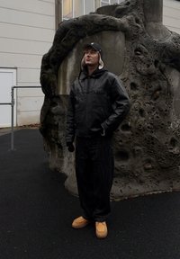 Jachetă de piele neagră, mănuși negre, blugi închiși la culoare și încălțăminte maro. Stă lângă o structură texturată asemănătoare unei stânci într-un decor exterior.