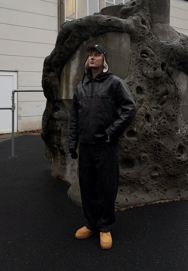 Giacca di pelle nera, guanti neri, jeans scuri e calzature marroni. In piedi vicino a una struttura rocciosa con texture in un ambiente all'aperto.