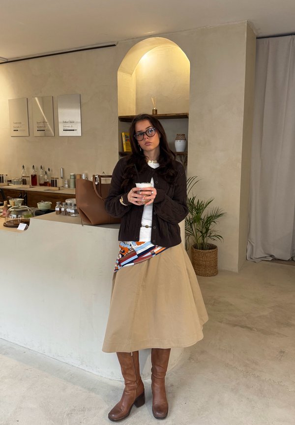 Ženska z očali in rjavim jopičem, ki drži skodelico, stoji v minimalistični kavarni z bež notranjo opremo, sobno rastlino in policami s knjigami ter kozarci.