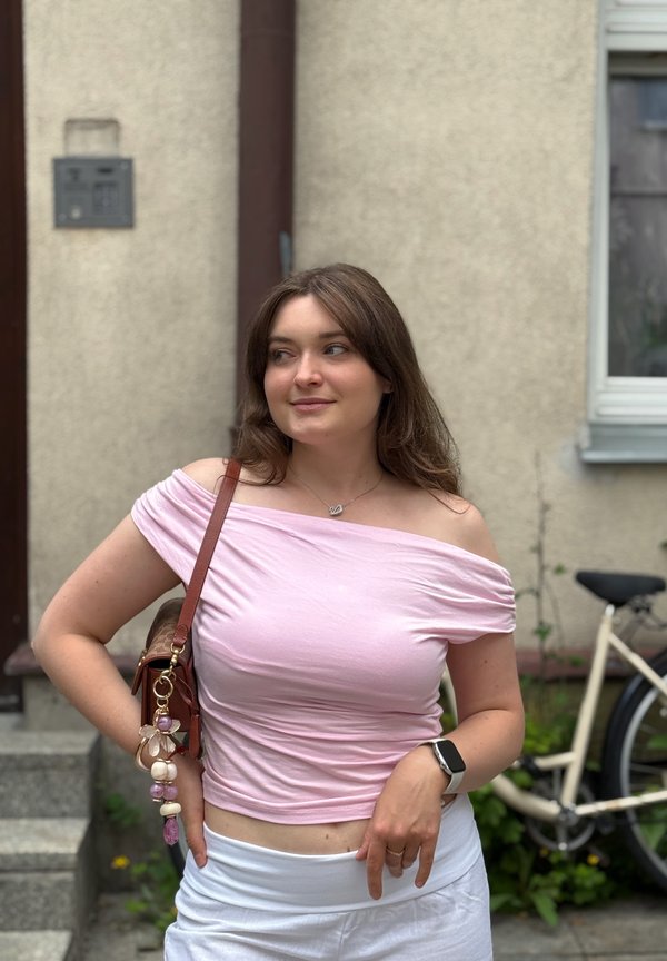 Ung kvinde iført lyserød off-shoulder top og hvide bukser står udendørs ved en cykel, bærende en brun skuldertaske og iført smartwatch.