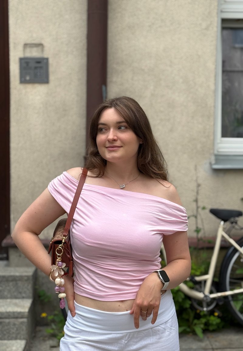Jeune femme portant un top rose épaules dénudées et un pantalon blanc, debout dehors près d'un vélo, portant un sac à bandoulière marron et une montre connectée.