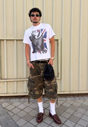 Joven con gafas de sol, camiseta blanca con gráfico, pantalones cortos cargo de camuflaje, calcetines blancos y zapatos marrones, sosteniendo una gorra negra junto a una pared metálica.