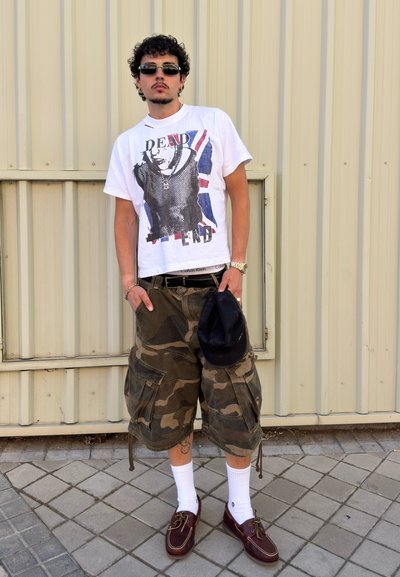 Joven con gafas de sol, camiseta blanca con gráfico, pantalones cortos cargo de camuflaje, calcetines blancos y zapatos marrones, sosteniendo una gorra negra junto a una pared metálica.