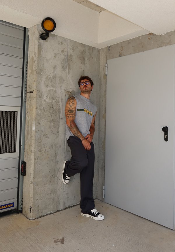Hombre con tatuajes, gafas, camiseta sin mangas y zapatillas apoyado contra una pared de hormigón cerca de una puerta gris cerrada y una puerta de garaje.