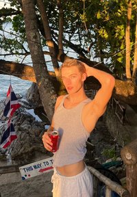 Persona, kas valkā pelēku bezrokas kreklu un baltus šortus, tur sarkanu kolas bundžu, stāv pie koka ar klinšu pludmali un karogiem fonā.