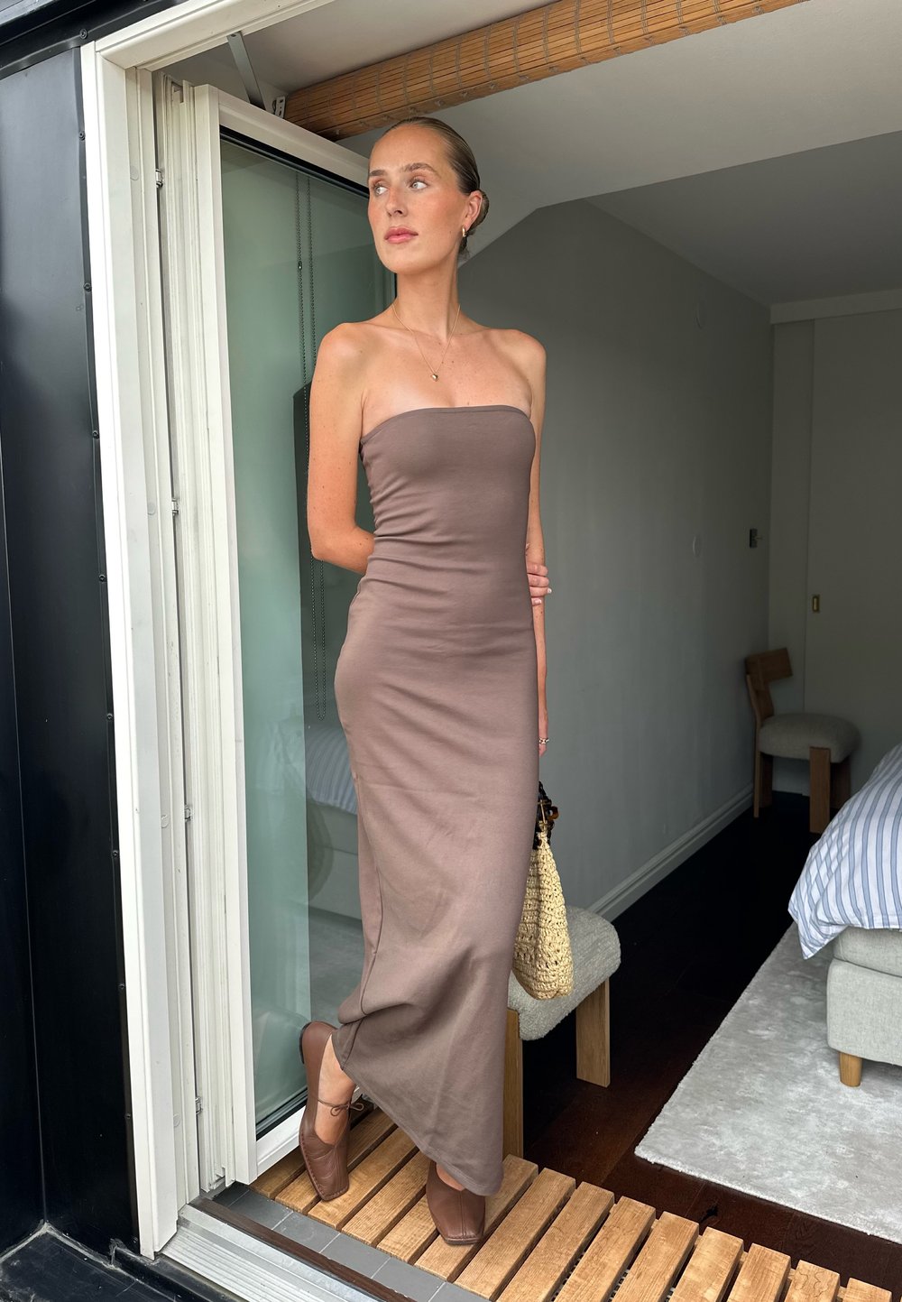 Brun axelbandslös maxiklänning, åtsittande design, mjukt tyg. Bärs med ljusbruna klackskor och en vävd väska. Står vid en glasdörr.