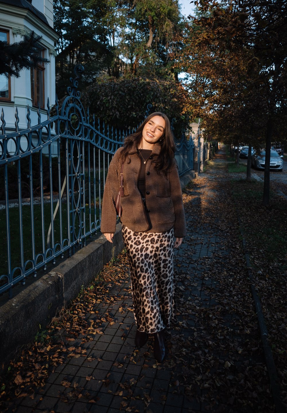 Woman smiling and walking on a leaf-covered sidewalk wearing a brown jacket and leopard-print skirt near iron fence and autumn trees.