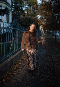 Woman smiling and walking on a leaf-covered sidewalk wearing a brown jacket and leopard-print skirt near iron fence and autumn trees.