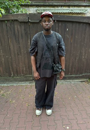 Chaleco acolchado negro sobre una camiseta gráfica negra, pantalones cargo negros holgados y zapatillas deportivas blancas gruesas. Mochila cruzada negra, de pie sobre un camino de ladrillos.