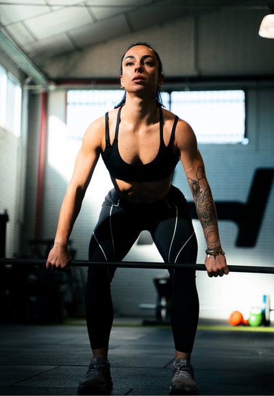 Mujer atlética con ropa deportiva negra agarrando una barra en un gimnasio, preparándose para levantar con una expresión concentrada y el brazo tatuado visible.