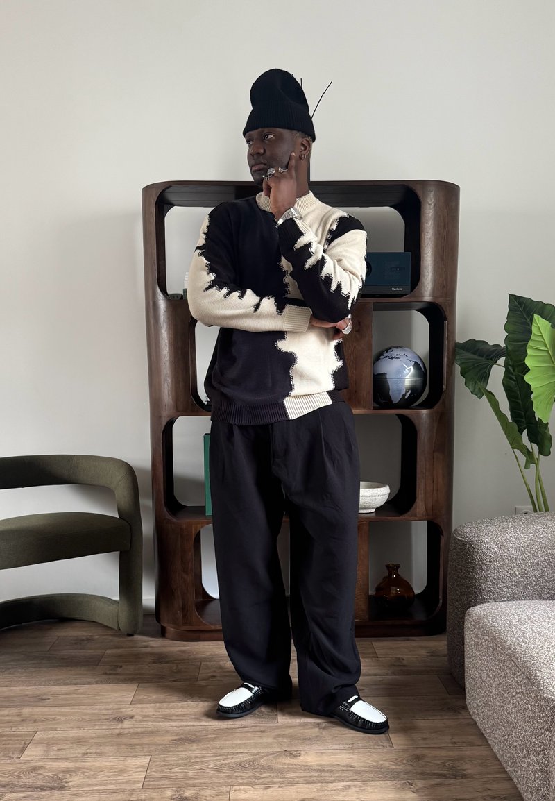 Άνδρας που φοράει μαύρο και λευκό πουλόβερ, μαύρο παντελόνι και μαύρο σκουφάκι, στέκεται σκυθρωπός σε ένα μοντέρνο σαλόνι με ξύλινο πάτωμα.