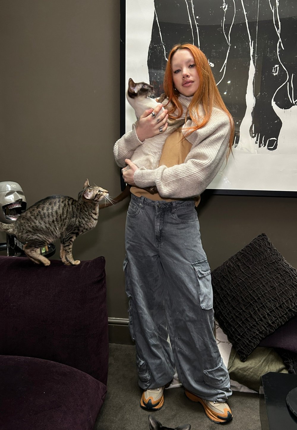 A young person with long reddish-orange hair is wearing a light sweater over a beige top while holding a white cat. Grey cargo trousers and patterned cushions are also visible.