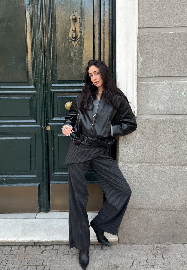 Moteris su ilgais tamsiais plaukais, apsirengusi juoda odine striuke ir dryžuotomis kelnėmis, atsirėmusi į tamsiai žalių durų su žalvarine rankena ir pilką sieną.