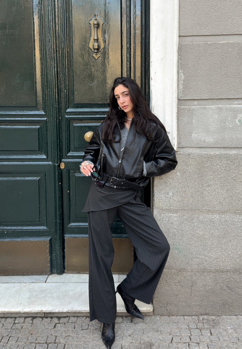Femme aux longs cheveux bruns portant une veste en cuir noire et un pantalon à rayures, appuyée contre une porte vert foncé avec une poignée en laiton et un mur gris.