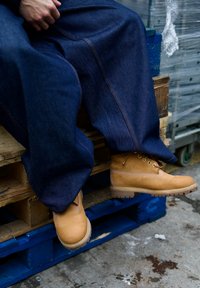 Personne portant des bottes de travail couleur beige et un pantalon en denim bleu foncé assise sur des palettes en bois empilées dans un environnement industriel.