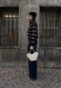 Jeune femme portant un pull rayé, un jean foncé, un béret noir, tenant un sac à main blanc, debout sur une rue pavée à côté d'un bâtiment en pierre avec des fenêtres grillagées.