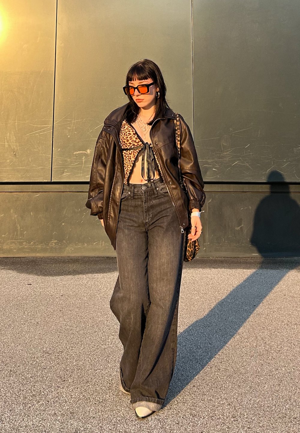 Veste en cuir noire oversize, crop top à imprimé léopard, jean large foncé, lunettes de soleil rectangulaires orange, et sac à main à motifs.