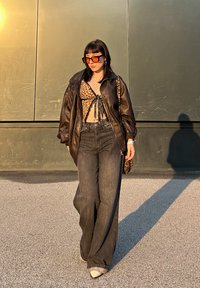 Veste en cuir noire oversize, crop top à imprimé léopard, jean large foncé, lunettes de soleil rectangulaires orange, et sac à main à motifs.