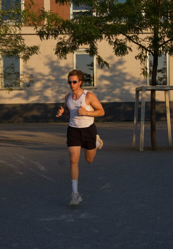 Personne portant des lunettes de soleil, un débardeur blanc et un short noir en train de courir dans une rue urbaine ensoleillée avec des arbres et des fenêtres de bâtiments en arrière-plan.