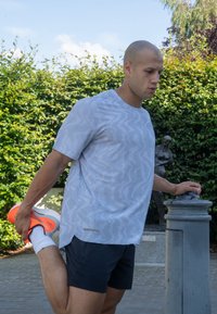 Hellgraues Sportshirt mit Wellenmustern, kurzen Ärmeln und seitlichen Schlitzen, kombiniert mit schwarzen Shorts und knallorangen Laufschuhen.