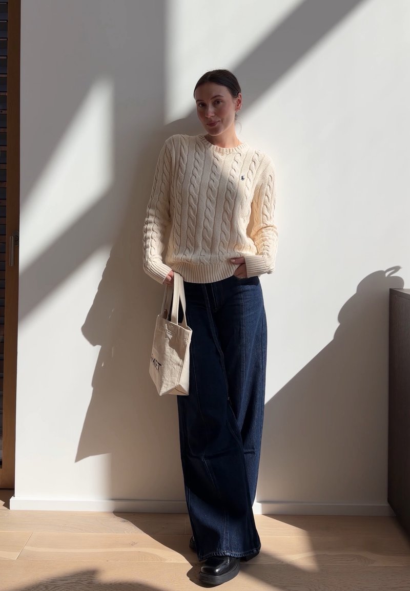 Femme debout à l'intérieur portant un pull torsadé crème, un jean large foncé, des chaussures noires et tenant un sac fourre-tout beige.