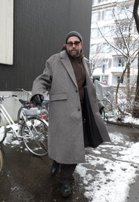 Homme barbu portant des lunettes de soleil, un long manteau gris, des gants noirs et un bonnet, marchant dans une rue urbaine enneigée près de vélos stationnés.