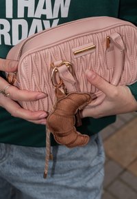 Růžová prošívaná kabelka s zlatými kovovými prvky a krátkými rukojeťmi. Obsahuje hnědý klíčenku ve tvaru croissantu. Nosená s casual džíny a zeleným topem.