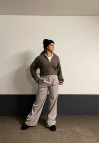 Sukienka z ribbowanego szarego materiału na zamek błyskawicznym, biała koszulka, jasnoszare spodnie z wysokim stanem i czarna czapka beanie; stojąca przed białą ścianą z ciemnym cokołem.