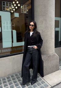 Femme portant un long manteau noir, un jean large noir, un cardigan noir, un haut blanc et des lunettes de soleil, debout contre une colonne en pierre près d'une fenêtre en verre.