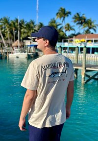 Homme portant un t-shirt de pêche beige "Marine Trophy" et une casquette marine, debout près d'une eau turquoise avec des bateaux et des palmiers en arrière-plan.