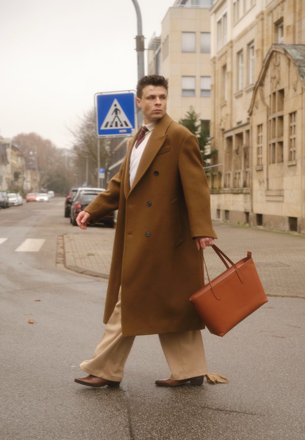 Palton maro cu nasturi dubli, pantaloni bej, pantofi maro, ținând o geantă tote din piele portocalie. Scenă stradală cu clădiri și semne de circulație în fundal.