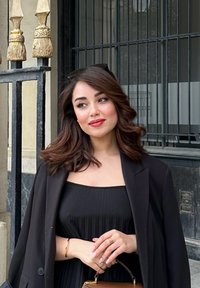 Frau in einem schwarzen, plissierten Kleid und schwarzem Blazer, die eine braune Handtasche hält. Lange, wellige Haare, sanftes Make-up, steht neben einem dekorativen Eisenzaun.