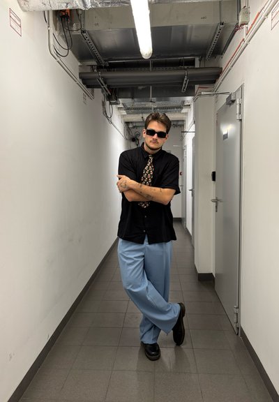 Camisa negra de manga corta, corbata a rayas, pantalones de pierna ancha de color azul claro y zapatos negros. Lleva gafas de sol, con los brazos cruzados, de pie en un pasillo.