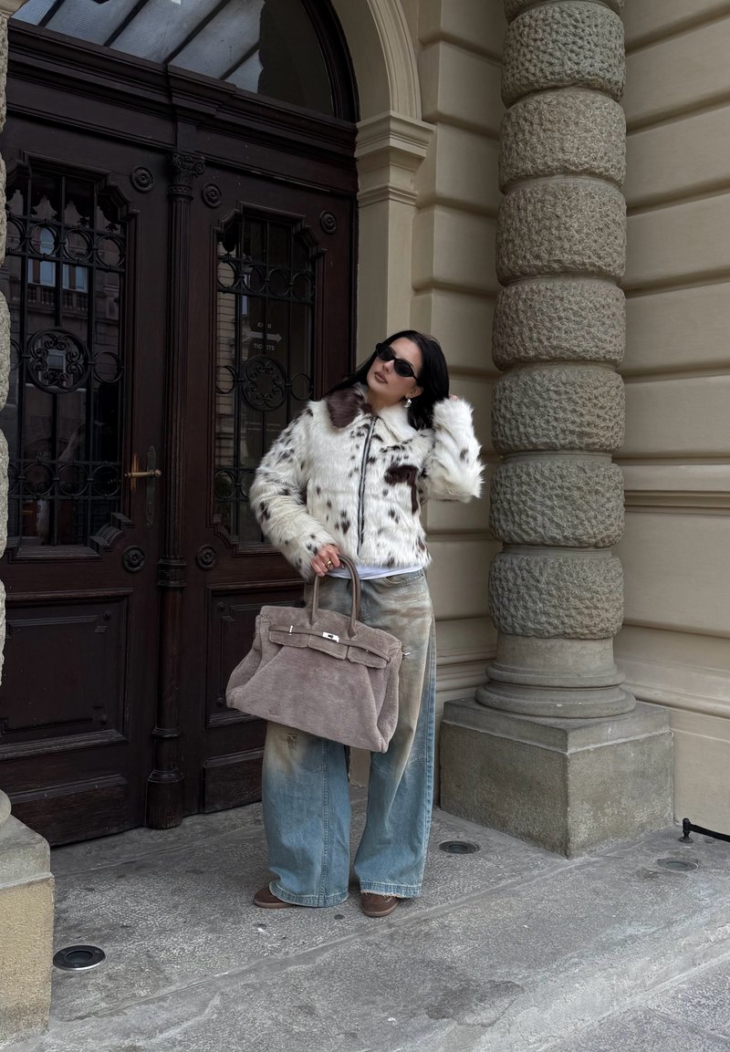 Donna con giacca di pelliccia maculata e jeans larghi che tiene in mano una grande borsa beige, in piedi accanto a una porta in legno scuro finemente intagliata e a una colonna in pietra testurizzata.