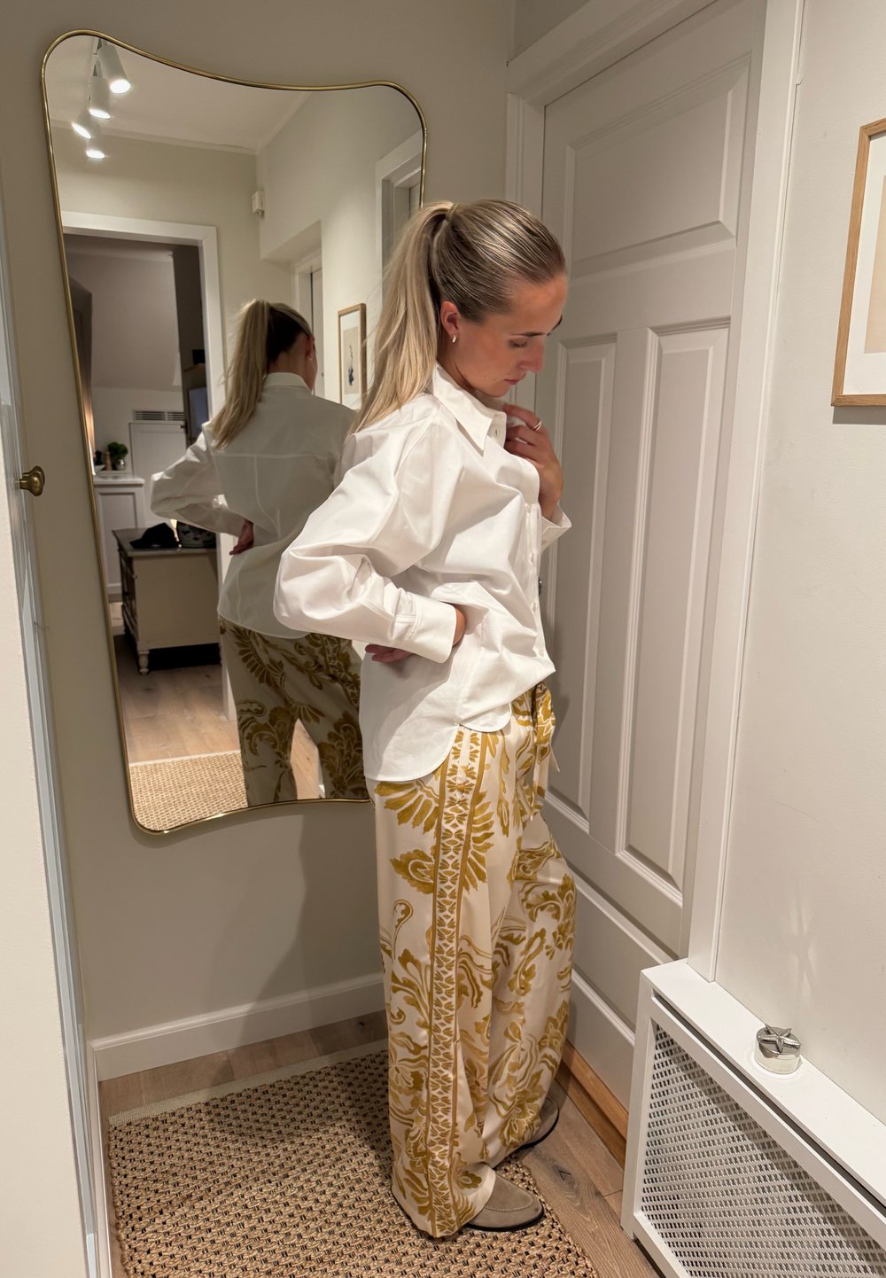 Camisa blanca de manga larga combinada con pantalones anchos crema con estampados florales dorados. Tela suave y ajuste relajado. Espejo de cuerpo entero al fondo.