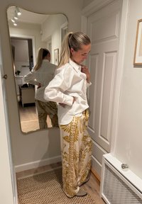 Camisa blanca de manga larga combinada con pantalones anchos crema con estampados florales dorados. Tela suave y ajuste relajado. Espejo de cuerpo entero al fondo.