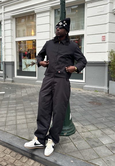 Chaqueta negra de nailon con cremallera en la parte delantera, pantalones negros holgados y zapatillas Nike blancas. Los accesorios incluyen un gorro negro con acentos decorativos.