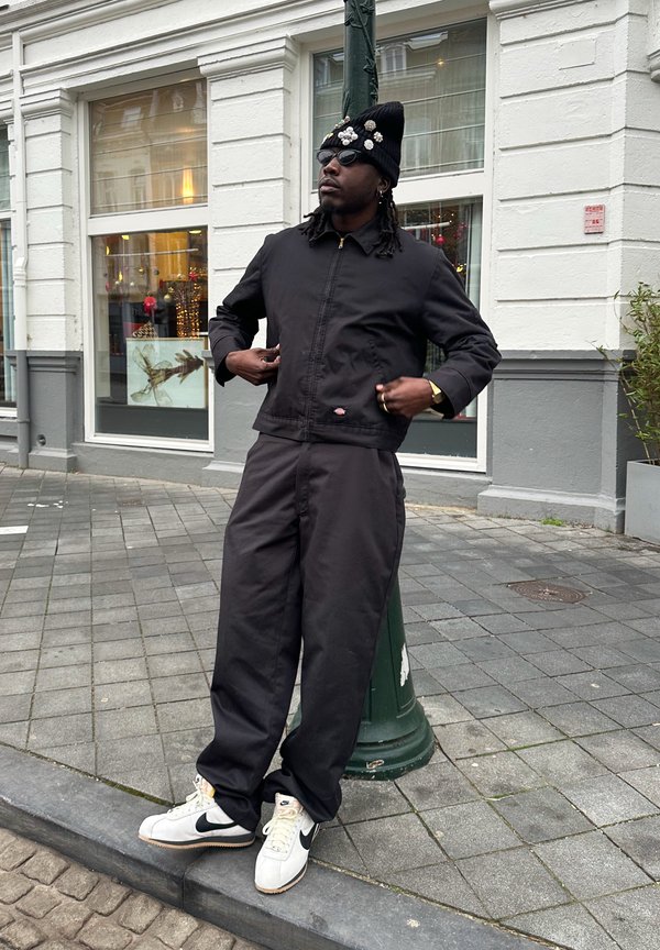 Casaco preto de nylon com fecho de correr, calças pretas largas e ténis brancos da Nike. Os acessórios incluem um gorro preto com detalhes decorativos.