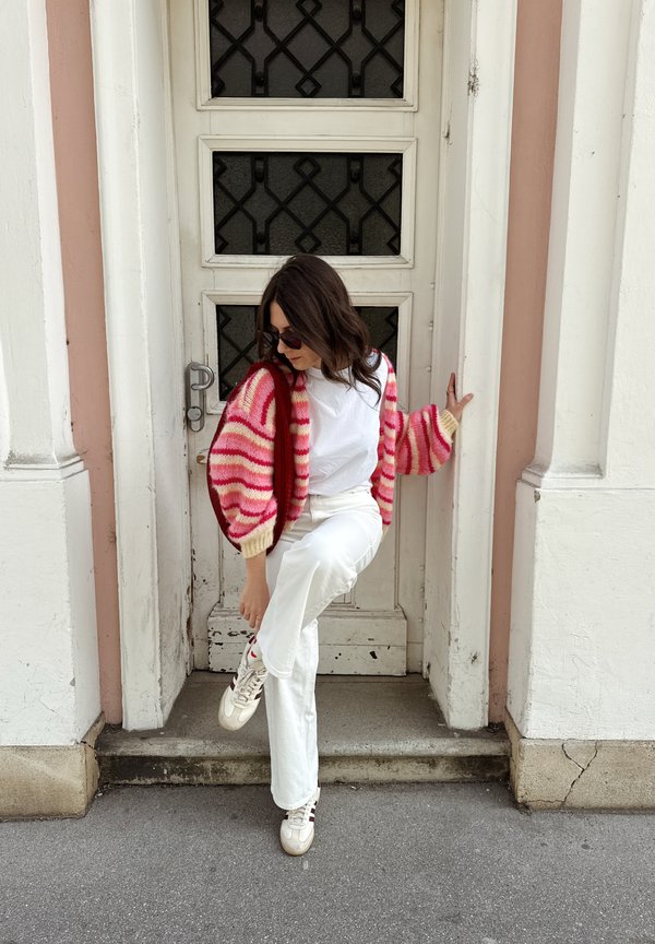 Femme en pantalon blanc, chemise blanche et cardigan rose à rayures ajustant sa chaussure en se tenant dans une porte avec des panneaux de fenêtres noirs décoratifs.