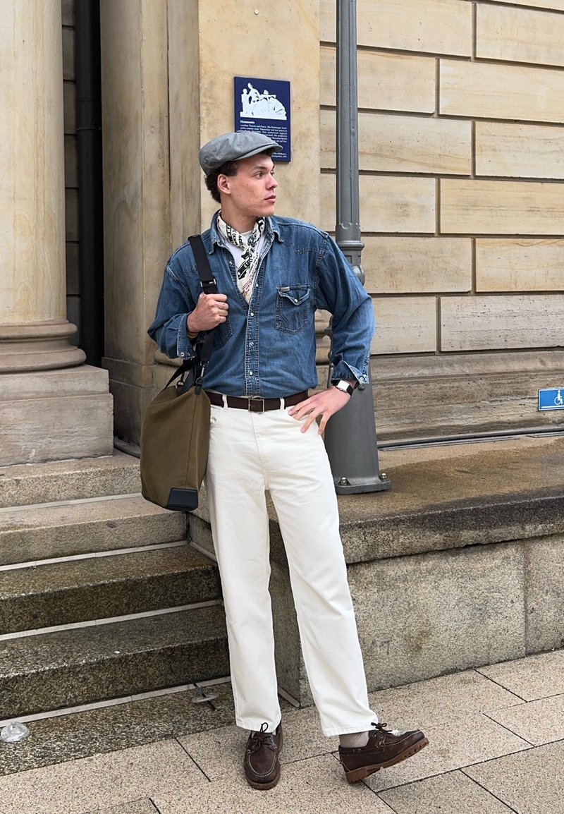 Man wearing denim shirt, white trousers, grey cap and scarf stands near stone steps outside a building carrying an olive bag on his shoulder.