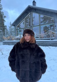 Jeune femme aux cheveux bouclés portant un manteau en fourrure sombre et un chapeau, debout dans la neige devant une cabane moderne décorée de guirlandes lumineuses.