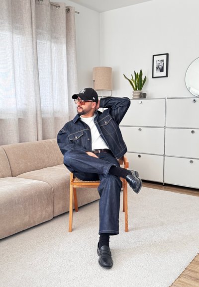 Hombre con chaqueta y pantalones vaqueros, gorra negra y gafas, sentado con las piernas cruzadas en una silla de madera en un salón luminoso con sofá beige y armario blanco.