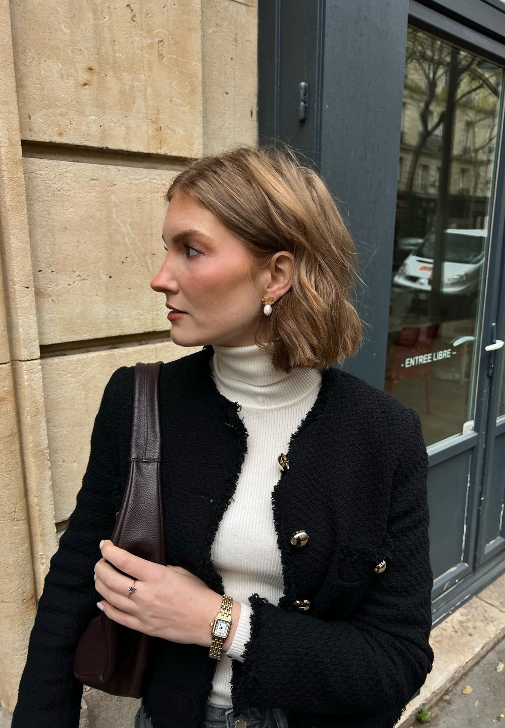 Zwarte blazer met textuur en gouden knopen, witte coltrui en een bruine schoudertas. Kenmerken zijn pareloorbellen en een gouden horloge om de pols.