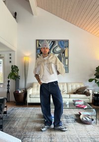 Een persoon staat met een grijze gebreide beanie, een wit T-shirt, een beige trui over de schouders gedrapeerd en losse blauwe denim jeans, met sneakers.
