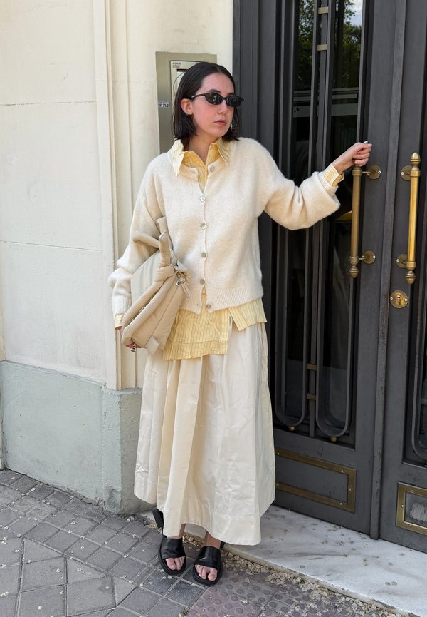 Kvinde iført en cremefarvet cardigan og nederdel holder dørhåndtaget med den ene hånd og en beige taske i den anden, iført solbriller og sorte sandaler.