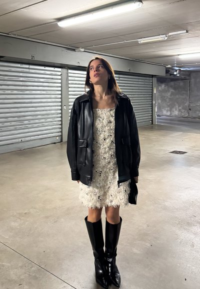 Mujer joven con un vestido blanco texturizado, chaqueta de cuero negra y botas negras hasta la rodilla, de pie en un estacionamiento de concreto.