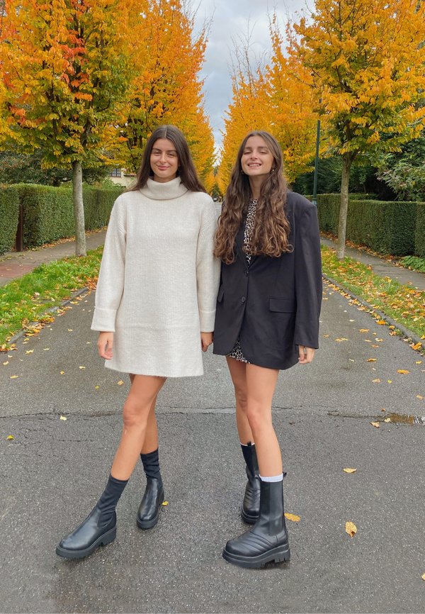 Dos mujeres de pie en una calle con árboles de otoño. Una lleva un vestido de suéter blanco y acanalado, la otra un blazer oversized negro con un vestido de leopardo debajo. Ambas tienen botas negras de tobillo.