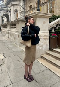 Svart oversized jakke, beige totebag, brun knelang skjørt, vinrøde sko med hæl og spenner, stående på en steinlagt fortau nær en bygning.